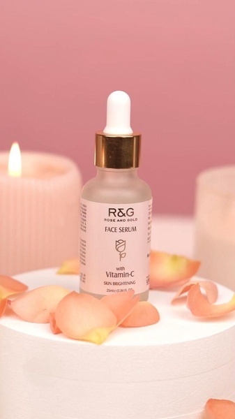 A high-end product hero shot of a face serum bottle placed on a white pedestal, surrounded by soft rose petals and a flickering candle against a muted pink background.